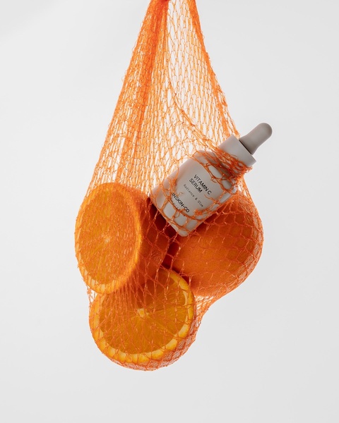 A high-contrast visual showcasing a Vitamin C serum bottle held within a bright orange mesh bag surrounded by fresh orange slices.
